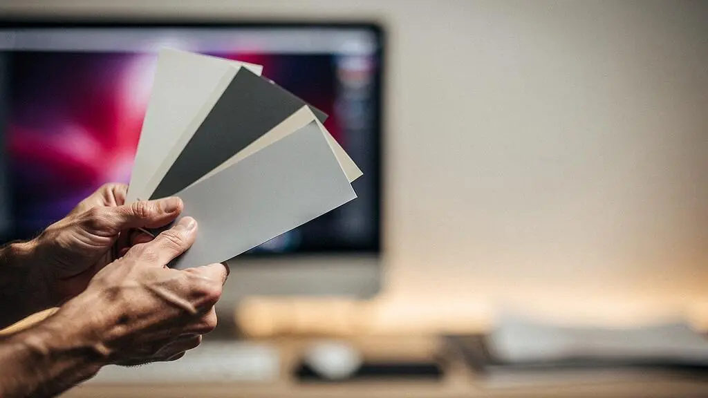 Mains de designer manipulant des nuanciers de couleurs papier avec en arriere-plan flou la lueur colorée d'un ecran, symbolisant la coherence entre print et web